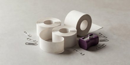 Soft-lit 3D render of curled thermal receipt rolls beside a plum-colored pencil sharpener and paper clips